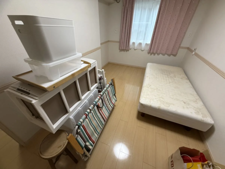 ピンクのカーテンがかかった小窓のある部屋。壁際にシングルの脚付きマットレスがあり、反対側には白い棚の上に収納ボックスやトレイが積み重なっている様子